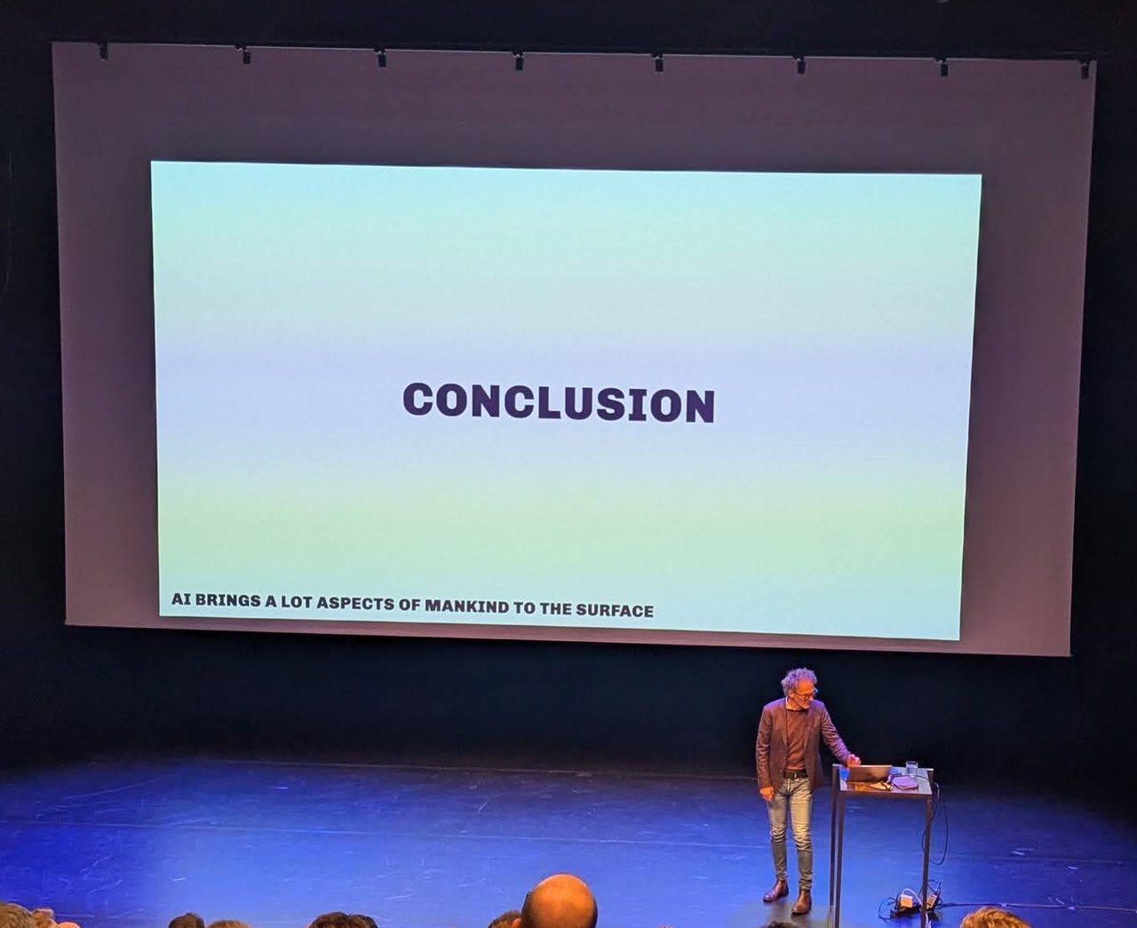 A HOPE 2025 speaker at the lectern in front of a large slide reading Conclusion, with the line AI brings a lot aspects of mankind to the surface