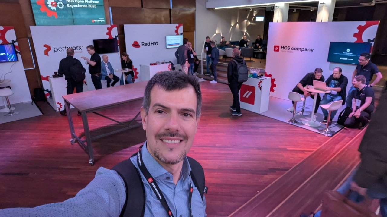 Selfie of Luca Berton on the HOPE 2025 expo floor with the Portworx, Red Hat and HCS company stands behind him and an HCS Open Platform Experience 2025 screen above