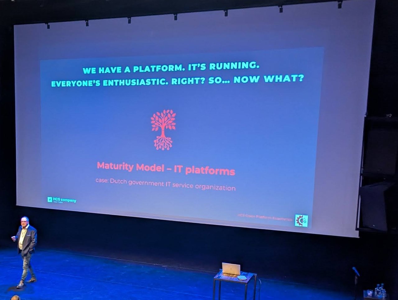 HOPE 2025 speaker on stage in front of a slide reading We have a platform. It's running. Everyone's enthusiastic. Right? So... now what? with the subtitle Maturity Model â IT platforms, case: Dutch government IT service organization