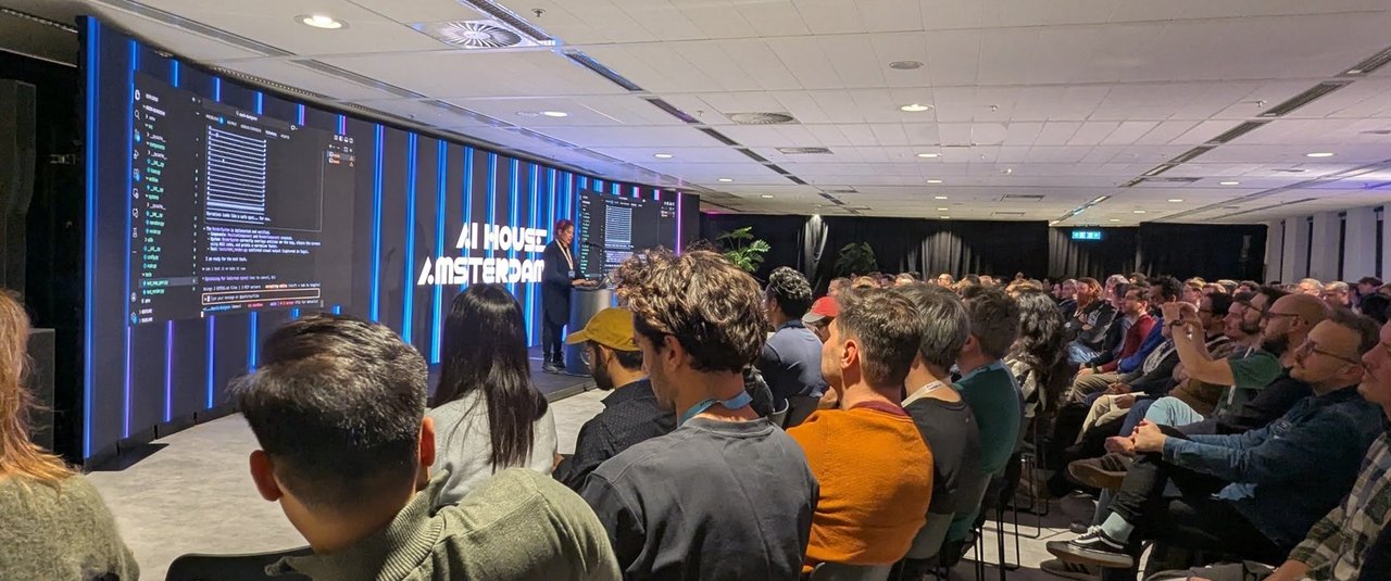 The speaker at the lectern with a terminal and code editor on the big screens while a seated audience watches the Gemini CLI demo