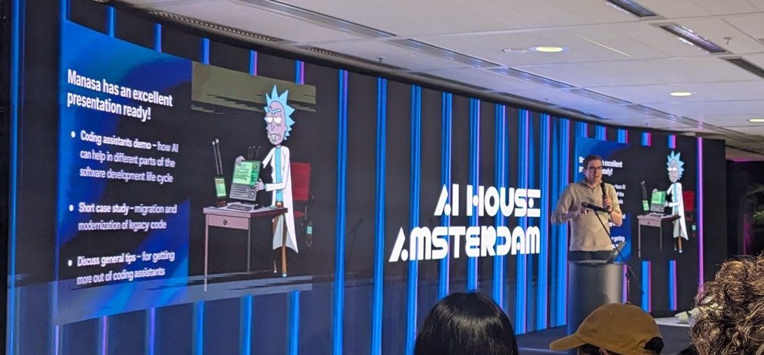 The AI House Amsterdam screen showing the session agenda with a cartoon scientist, while an organiser holds a microphone
