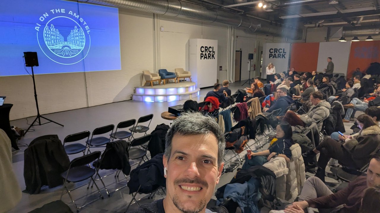 Luca Berton in the CRCL Park theatre before the AI on the Amstel robotics meetup, with the AI on the Amstel logo on screen and the empty panel chairs