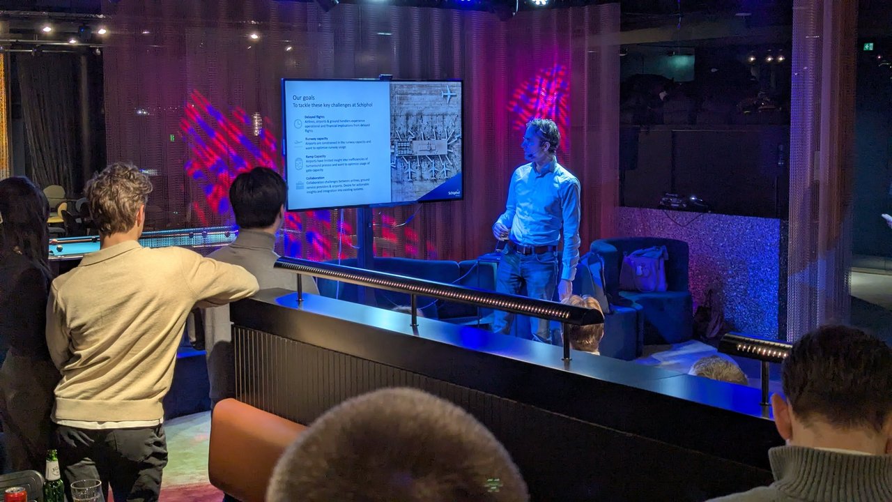 Speaker at AI Salon Amsterdam presenting the Our goals slide listing delayed flights, runway capacity, ramp capacity and collaboration at Schiphol