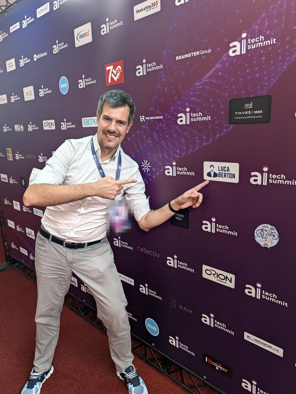 Luca Berton pointing at the LUCA BERTON speaker card on the AI Tech Summit sponsor wall, surrounded by sponsor and partner logos