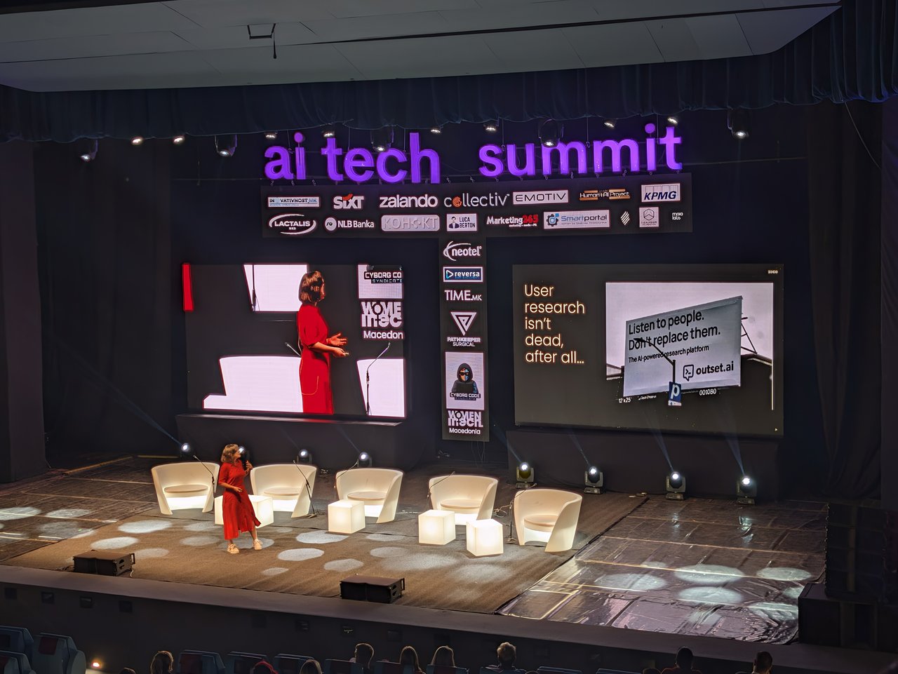 On the Main Stage, a speaker in a red dress next to a slide reading User research isn't dead, after all..., with a billboard photo that says Listen to people. Don't replace them.