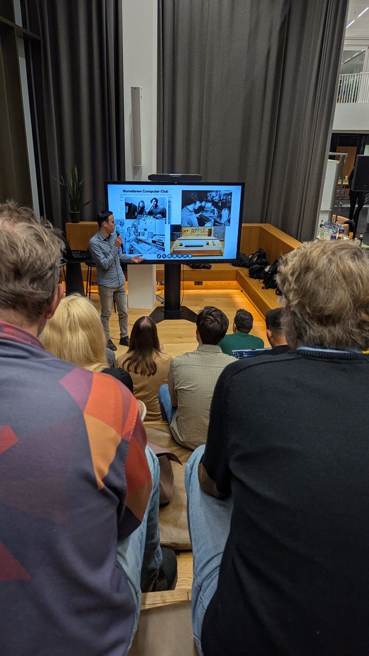 The host presenting a Homebrew Computer Club slide with archive photos and an Apple I, with the audience in front