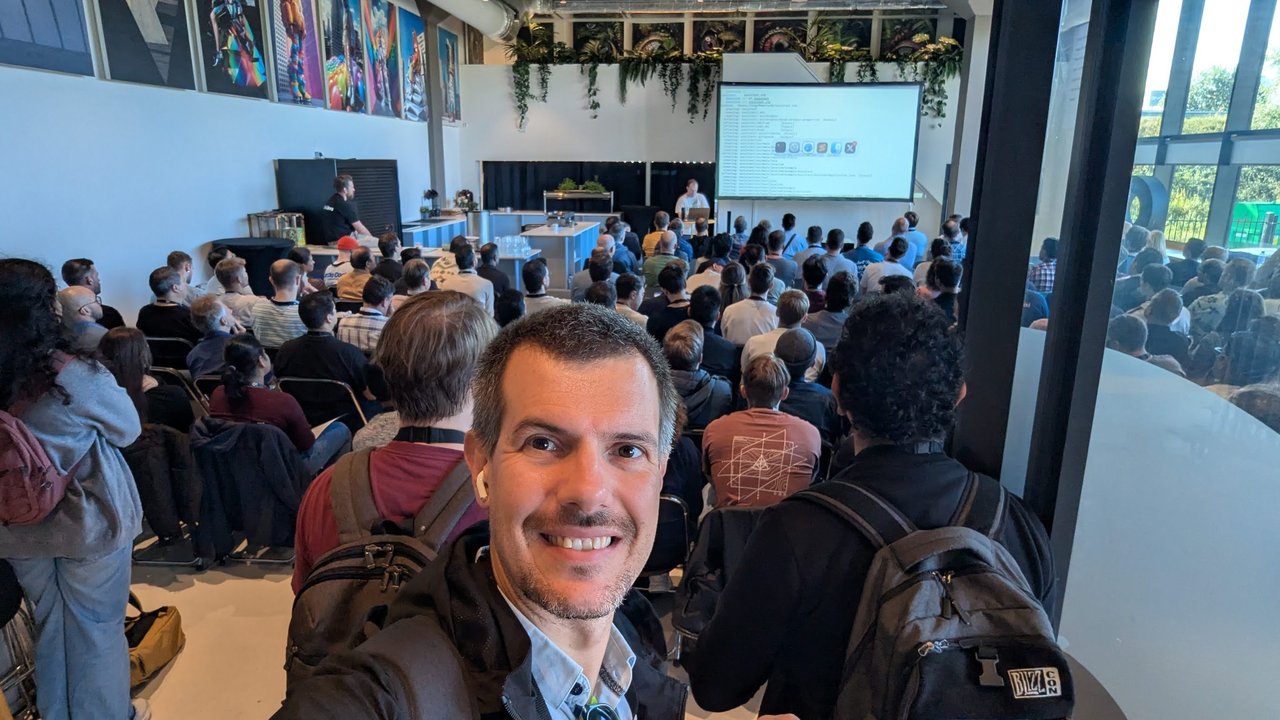 Luca Berton taking a selfie at the back of a packed keynote room at AI4Devs Amsterdam 2025, with a terminal on the projector screen