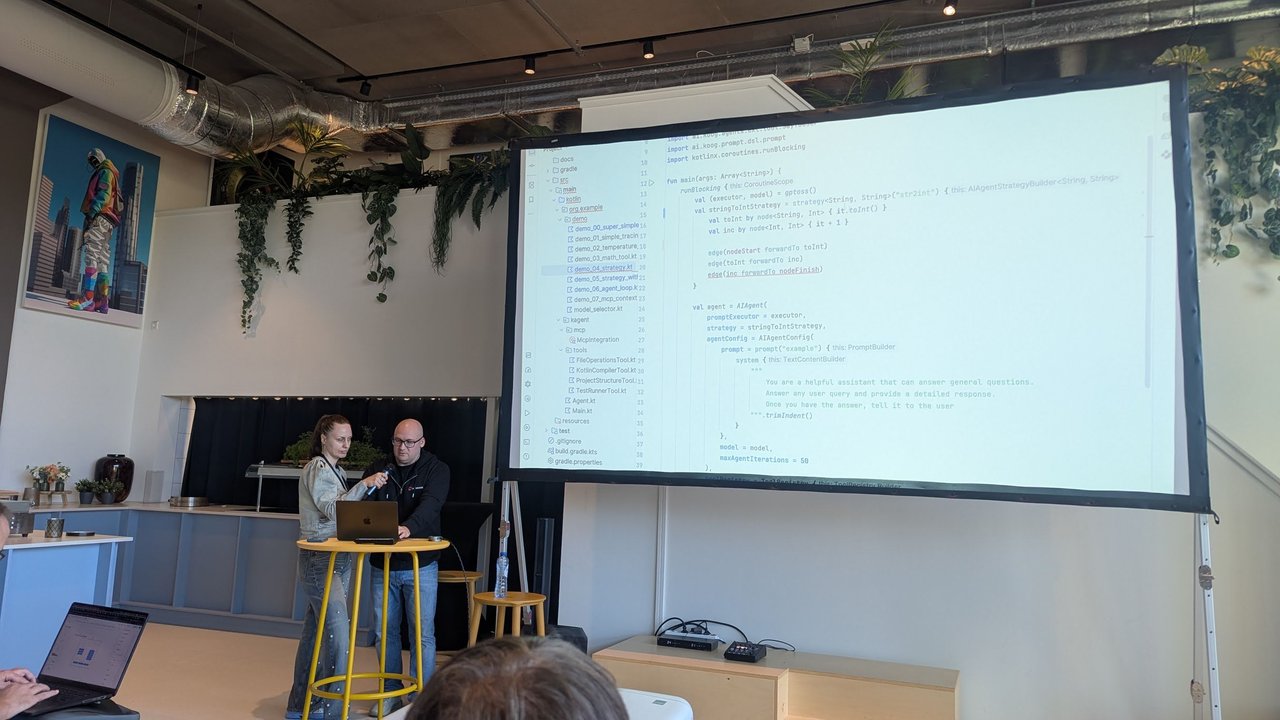 Two presenters at a high table with a laptop, next to a large screen showing Kotlin code that defines a Koog strategy with nodes toInt and inc and edges from nodeStart to nodeFinish