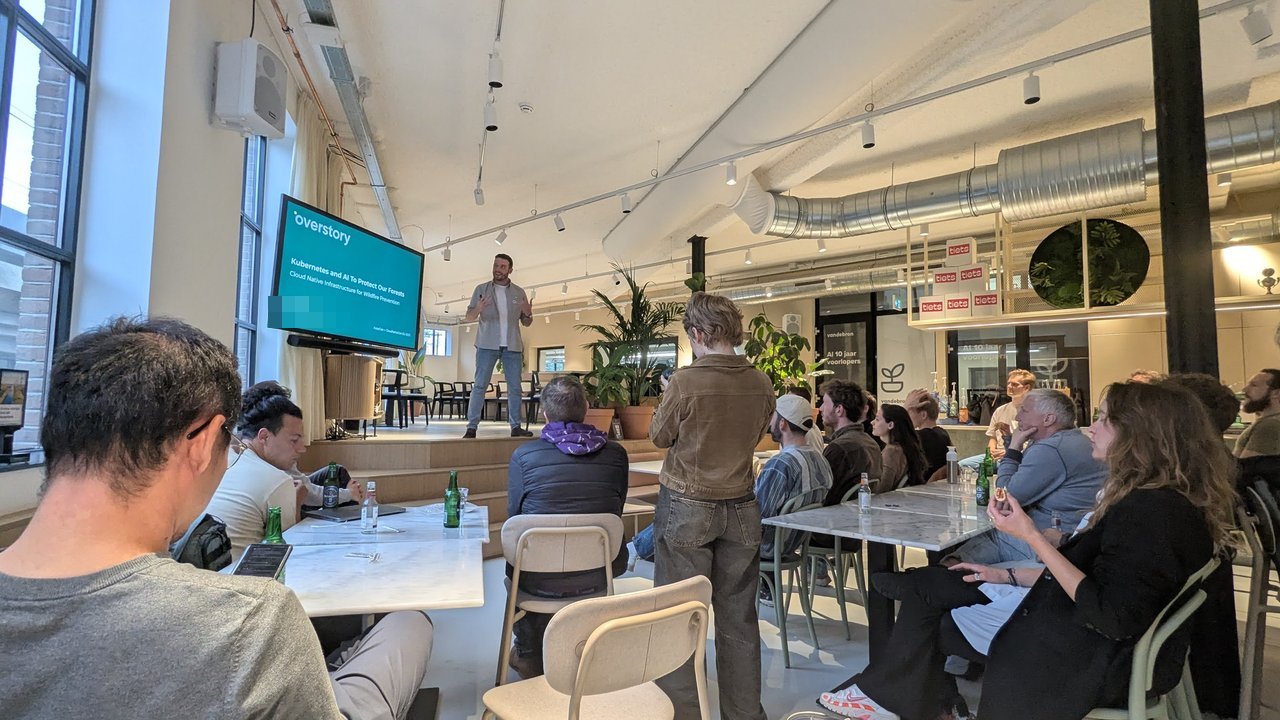 The Overstory title slide Kubernetes and AI To Protect Our Forests on a screen, with the speaker mid-gesture and attendees seated at tables