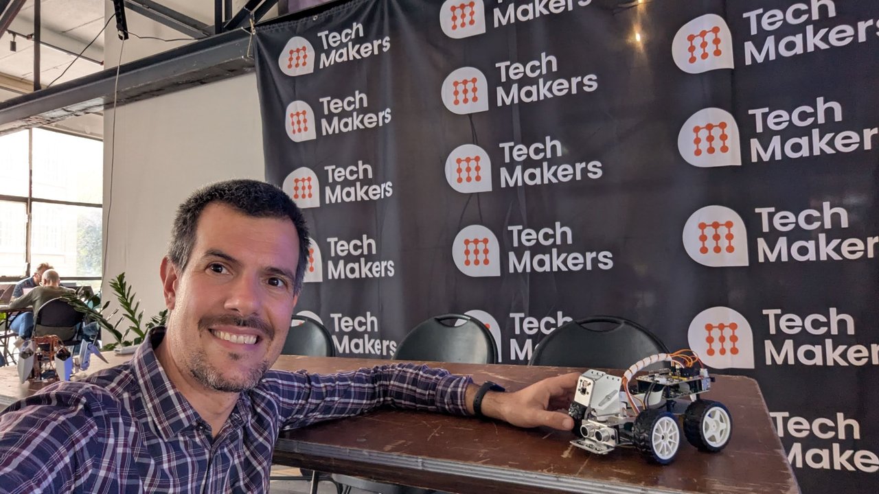 Luca Berton at the Tech Makers Top Bot Hackathon in front of a Tech Makers backdrop, with a small PiCar-X robot car on the table