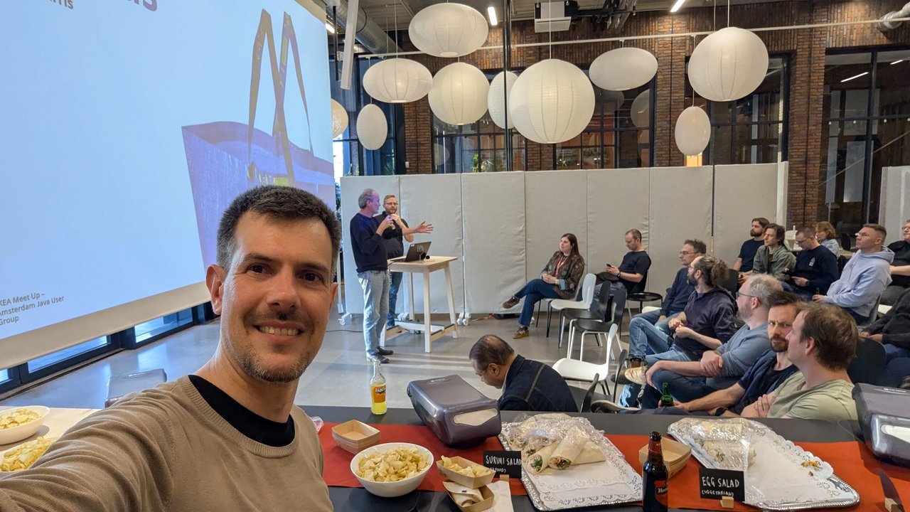 Luca Berton taking a selfie at the IKEA Meet Up of the Amsterdam Java User Group, with the hosts on stage, the audience and the food table behind him