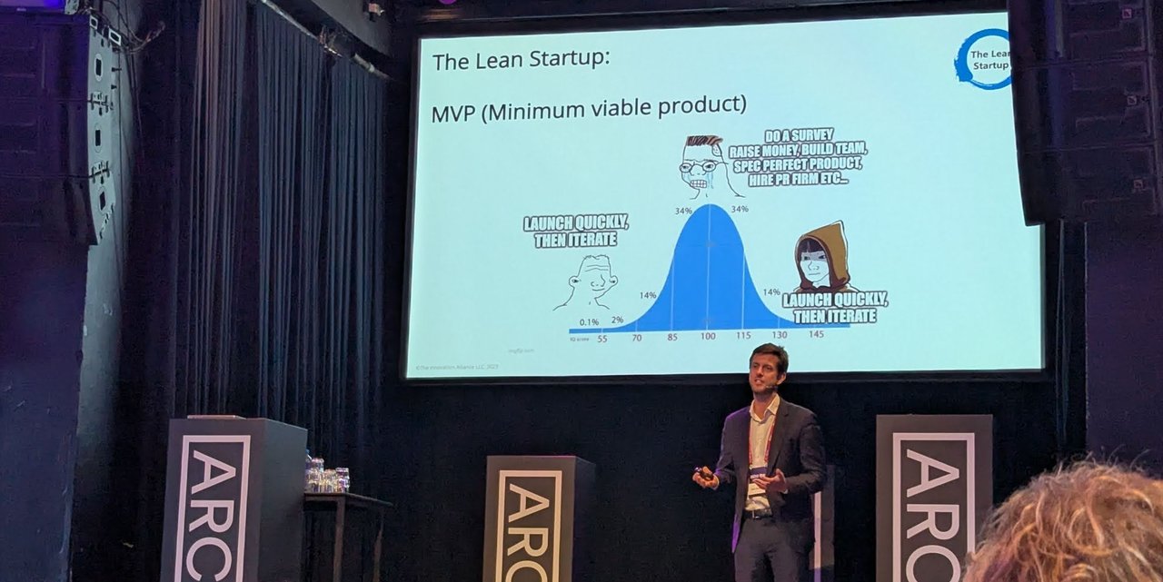Speaker on the Arch9 stage next to the slide The Lean Startup: MVP (Minimum viable product), a bell-curve meme where both ends say launch quickly, then iterate