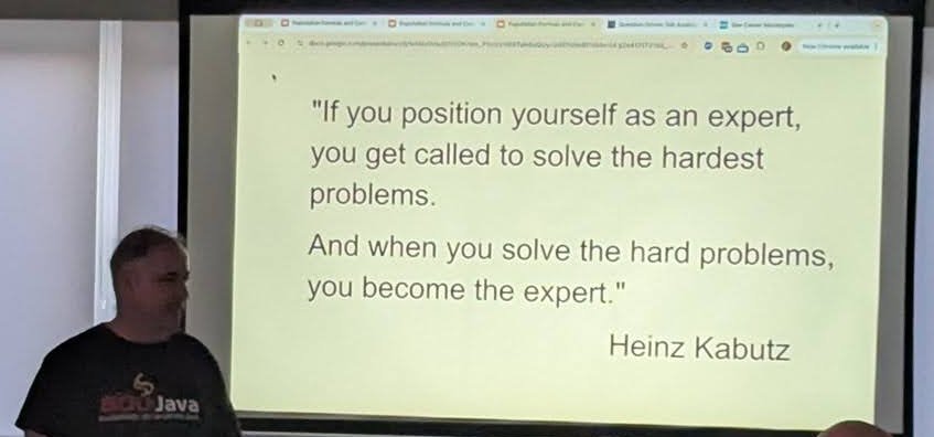 A speaker standing next to a slide with a quote attributed to Heinz Kabutz about positioning yourself as an expert