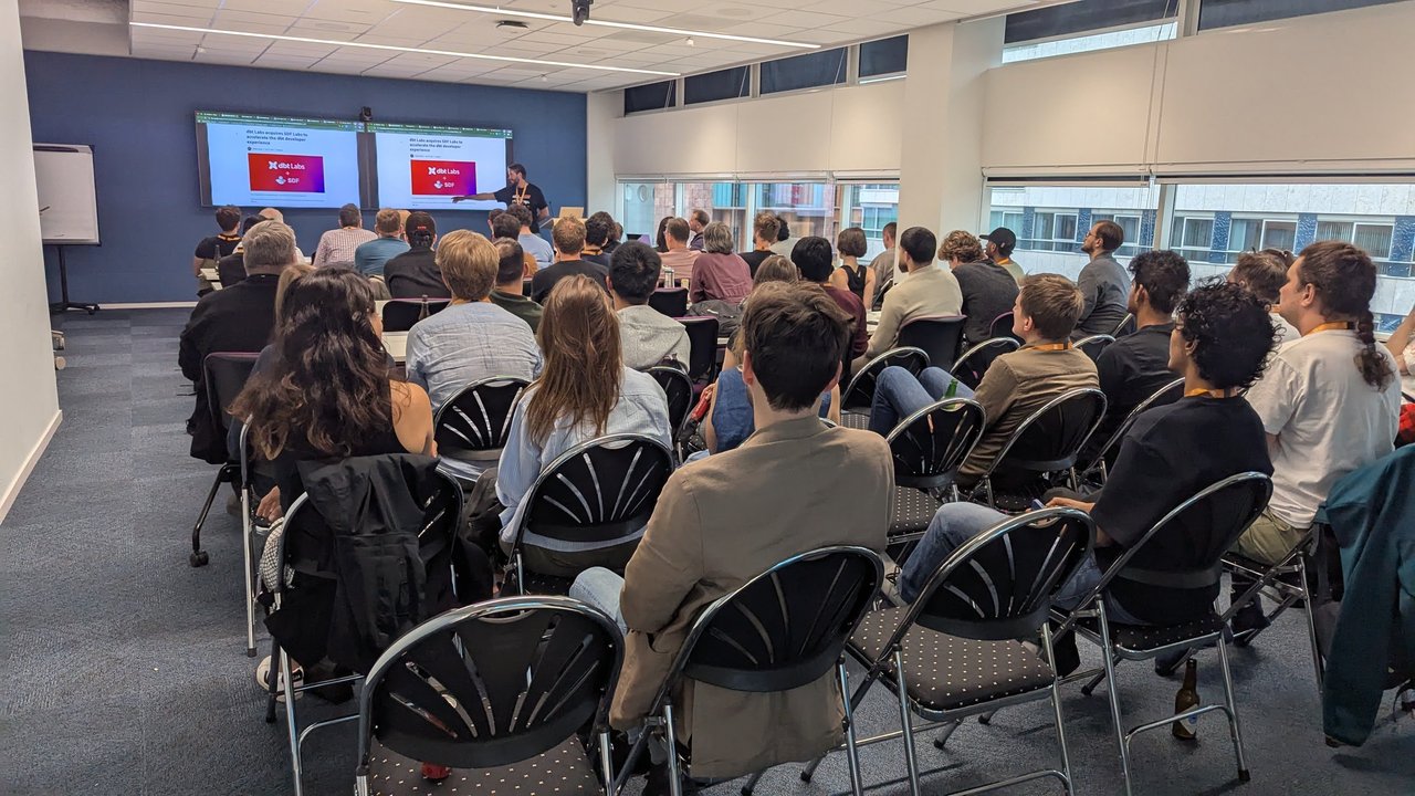 Meeting room full of attendees seen from behind, facing two screens showing the slide dbt Labs acquires SDF Labs to accelerate the dbt developer experience