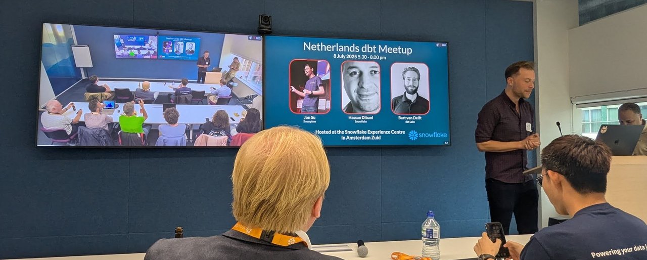 Two wall screens at the Snowflake Experience Centre, one showing a room camera view and one showing the Netherlands dbt Meetup title slide with three speakers and the Snowflake logo