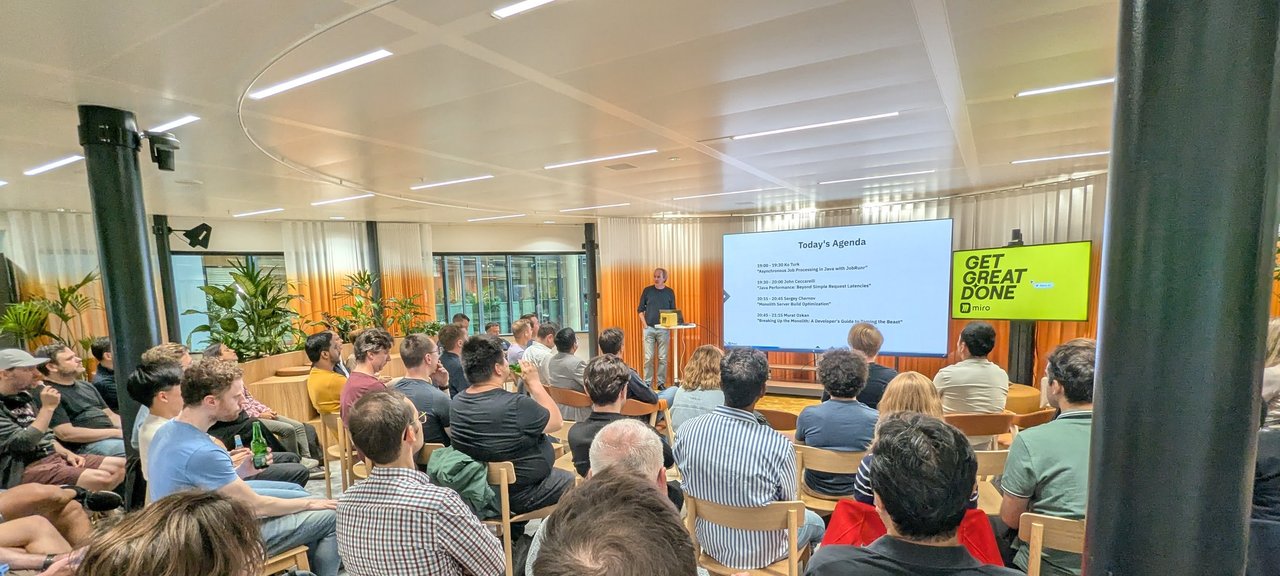 Wide view of the Amsterdam JUG audience facing a Today's Agenda slide listing four Java talks, with a Miro Get Great Done screen to the right