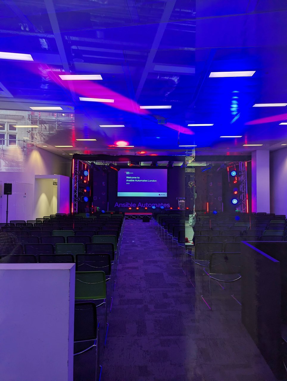 The main CodeNode hall in purple light before the doors opened, rows of empty chairs facing a screen that reads Welcome to Ansible Automates London 2023