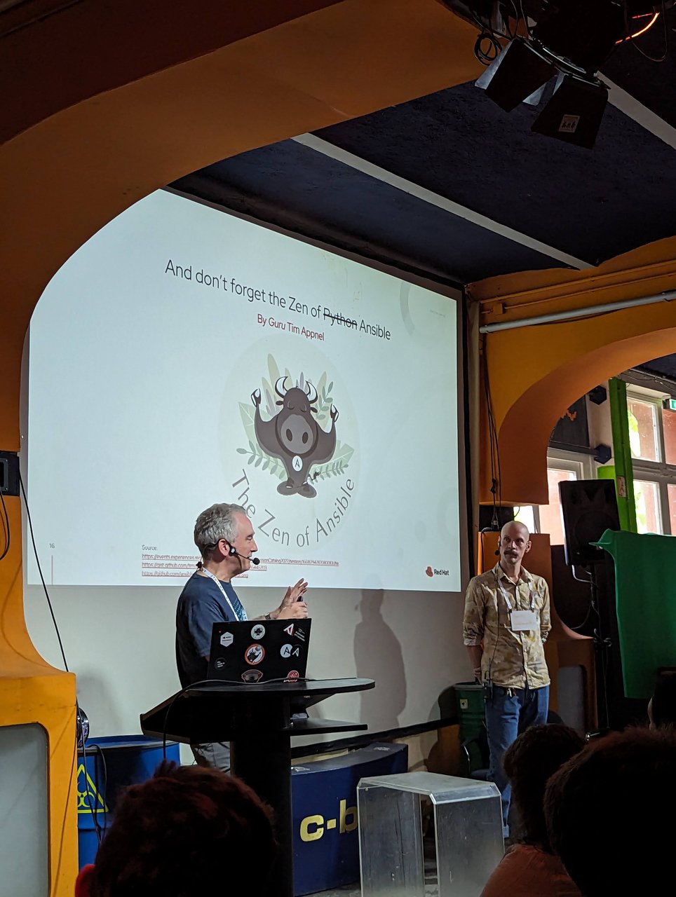 A slide at c-base reading "And don't forget the Zen of Python Ansible, by Guru Tim Appnel", with the two presenters on stage