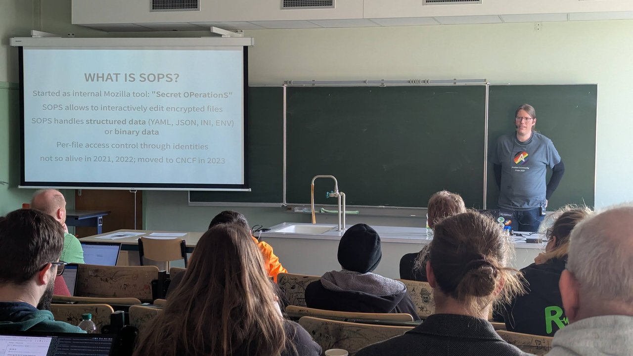 A "What is SOPS?" slide in a classroom at CfgMgmtCamp 2025 in Ghent, listing its Mozilla origins, interactive editing of encrypted files, structured and binary data, per-file access control and the 2023 move to the CNCF