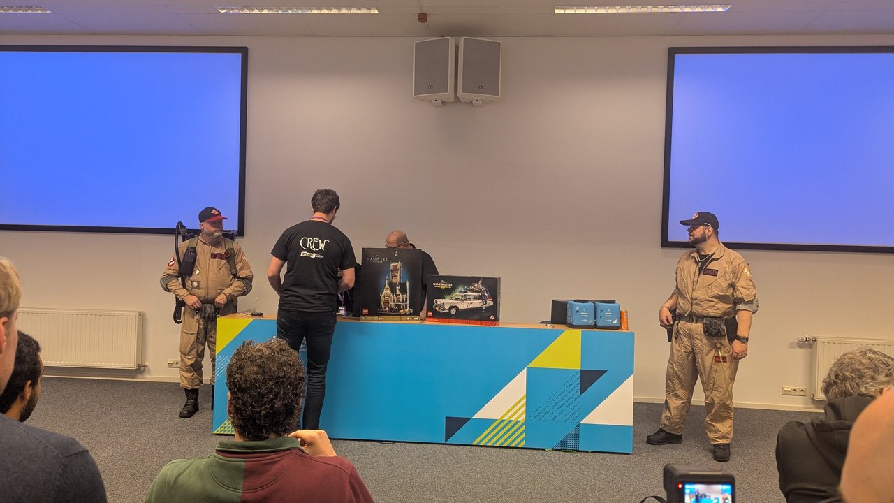 Two people in Ghostbusters costumes and a crew member at the stage table with LEGO Haunted House and Ghostbusters ECTO-1 boxes at BitBash 2025