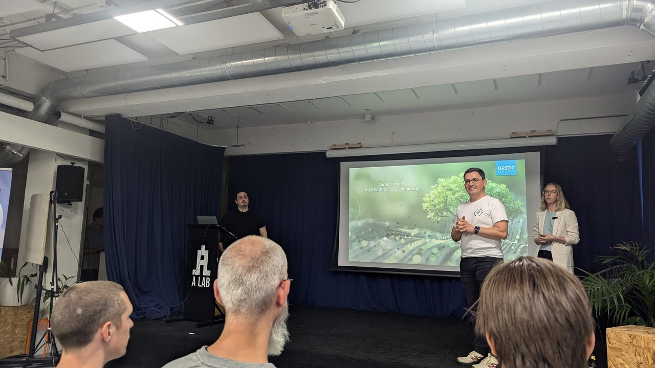 Two presenters on a small stage in front of a projected slide, with the A LAB sign beside the stage