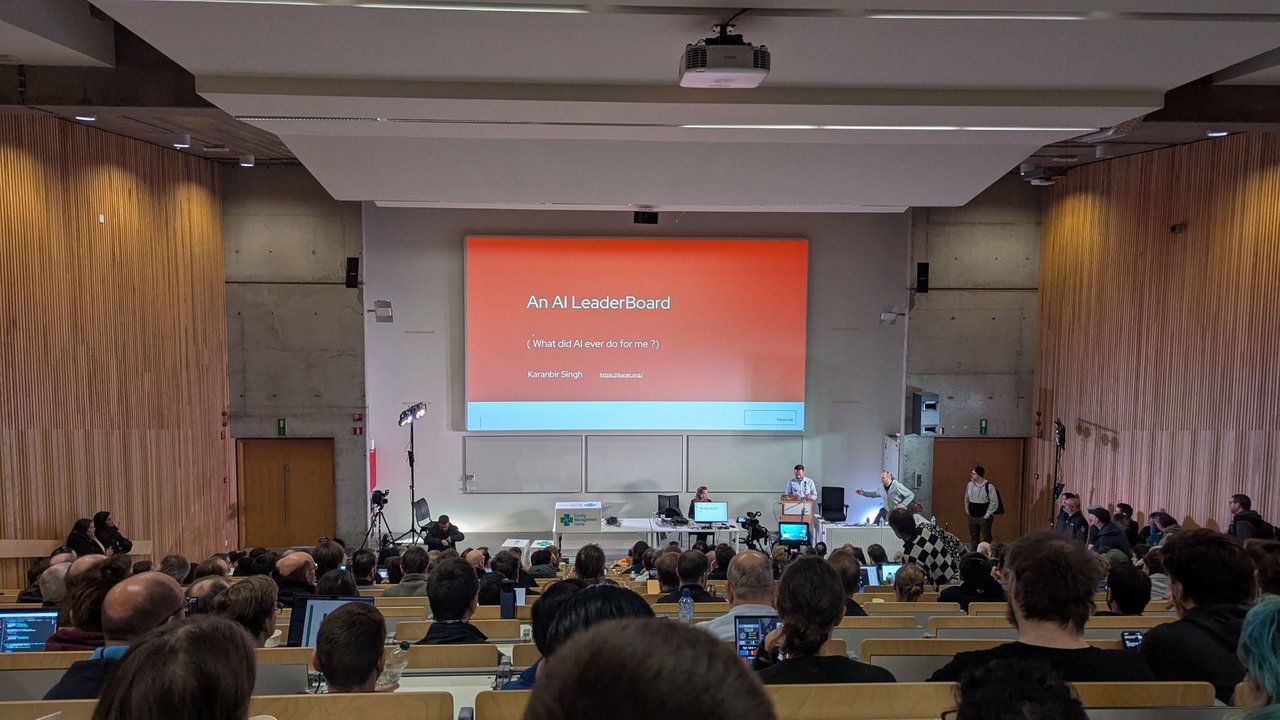 The title slide An AI LeaderBoard, What did AI ever do for me, by Karanbir Singh, in front of a full lecture hall at CfgMgmtCamp 2025