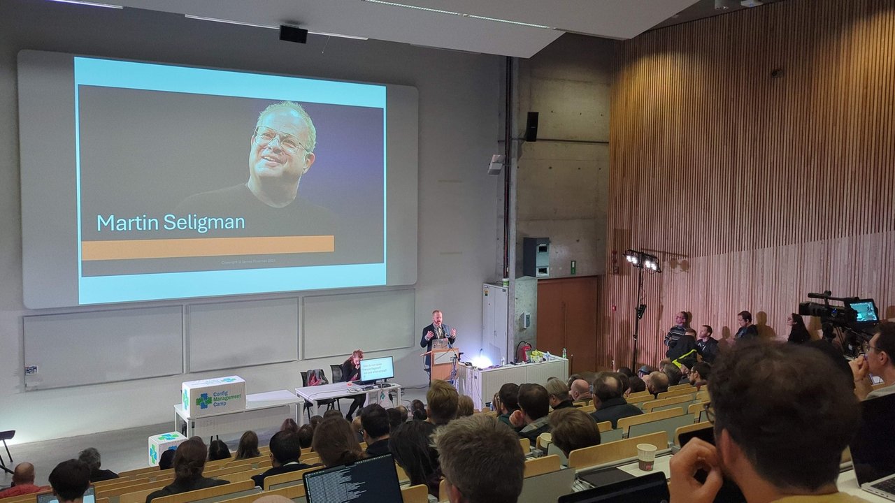 James Freeman on stage in the main auditorium at CfgMgmtCamp 2025, with a slide introducing Martin Seligman
