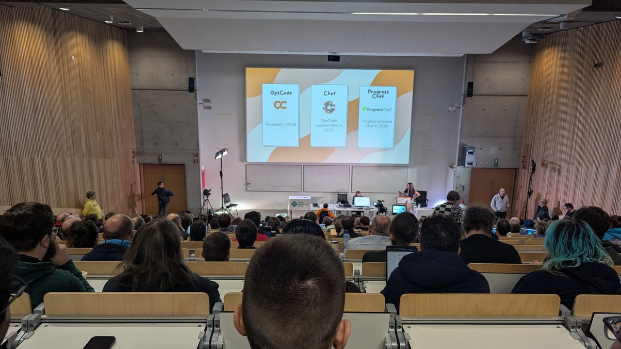 The OpsCode, Chef and Progress Chef timeline on screen in a full CfgMgmtCamp 2025 lecture hall in Ghent