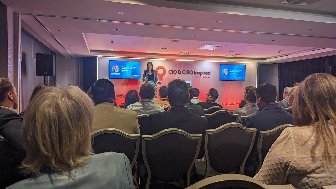 Speaker on stage between two screens showing the title Navigating the AI Frontier, Strategies for Effective Leadership, with the audience seen from behind