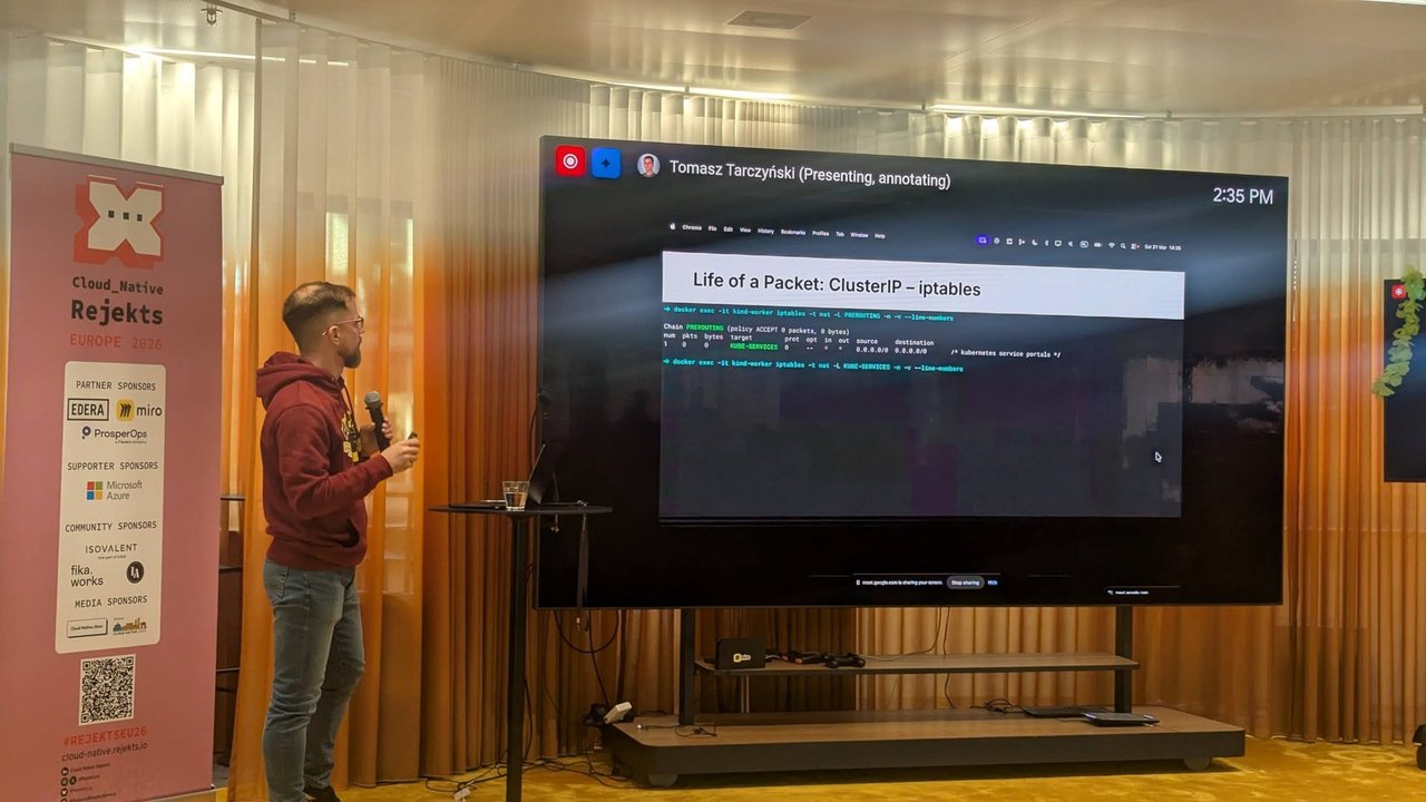 Tomasz Tarczyński presenting a Life of a Packet: ClusterIP iptables slide at Cloud Native Rejekts 2026, next to the Rejekts sponsor banner