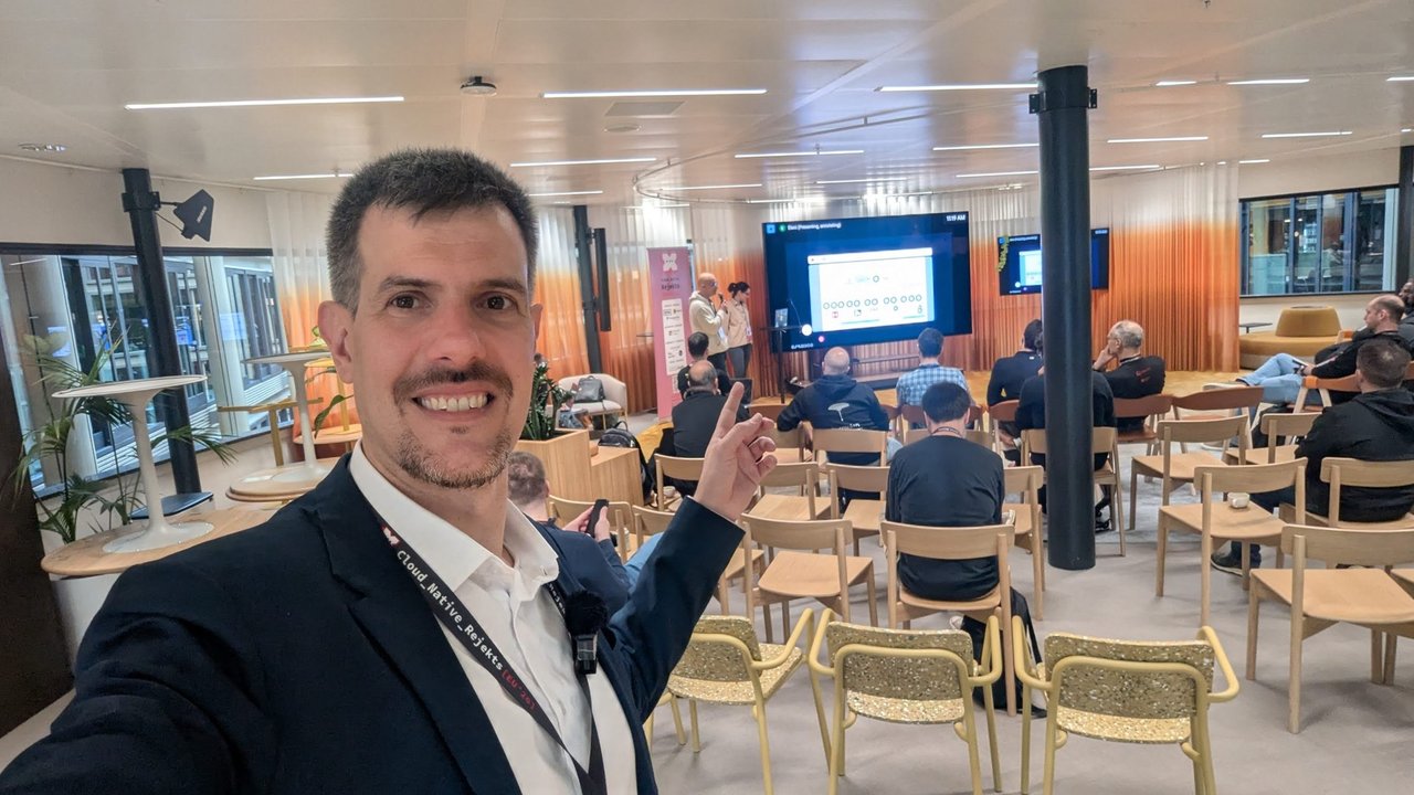 Luca Berton taking a selfie in Room 2 at Cloud Native Rejekts 2026, pointing at two presenters in front of the screen while the audience watches