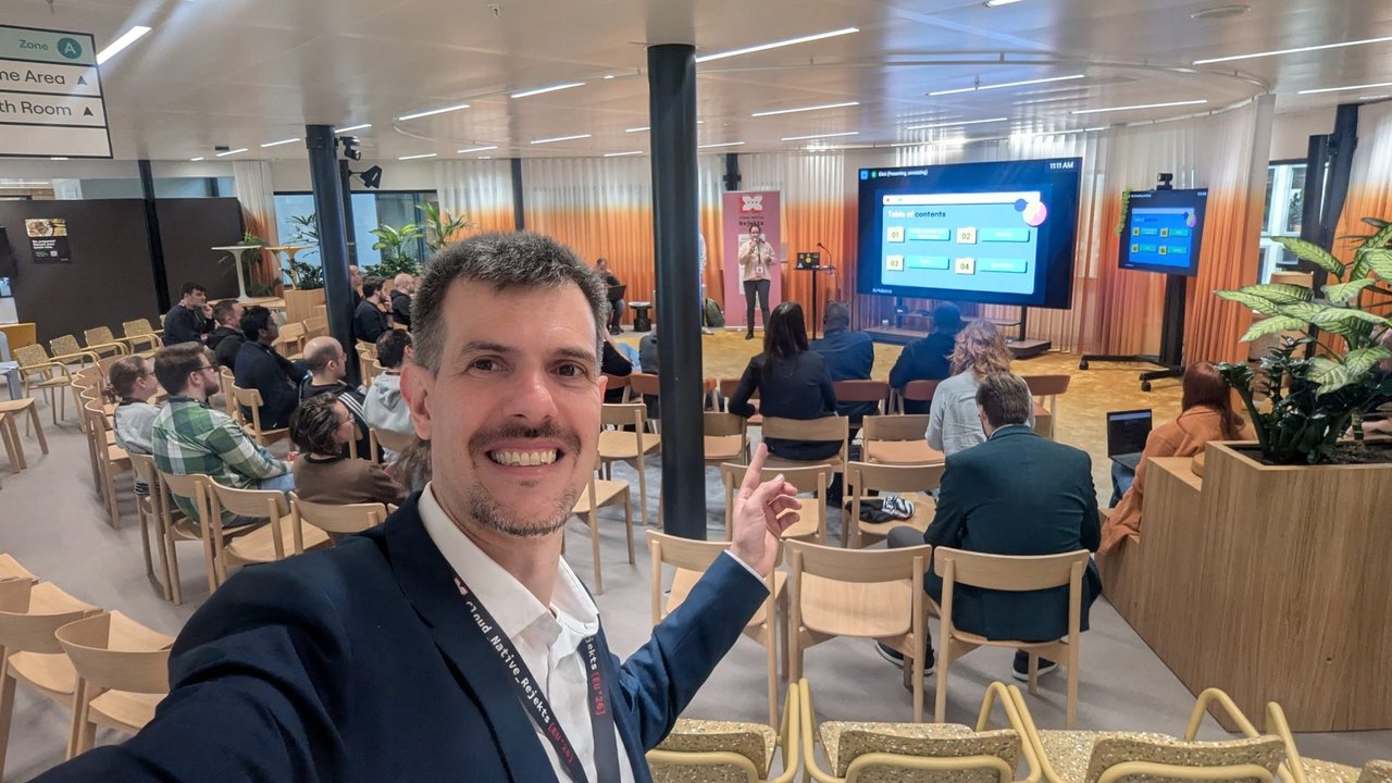 Luca Berton taking a selfie in the second room at Cloud Native Rejekts 2026 at the Miro office, pointing at the speaker and the screen behind the audience