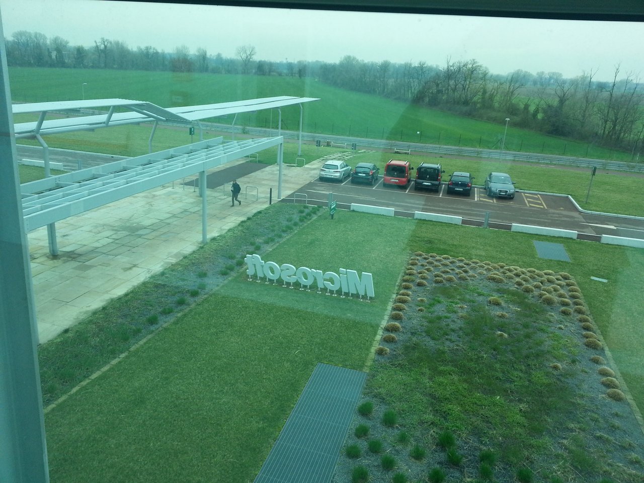 View through a window of the Microsoft Italia building in Peschiera Borromeo: white Microsoft letters standing on the lawn, a covered walkway and a car park