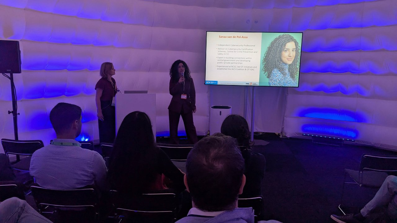 Sanaa van de Pol-Assa speaking with a microphone next to her introduction slide, which lists independent cybersecurity professional, advisor on cybersecurity certification schemes at the CCV, and experience at the NCSC leading OT initiatives
