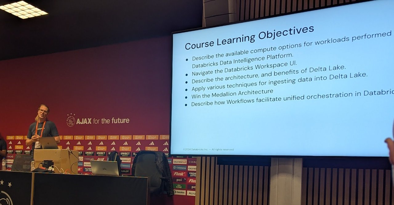 The instructor at the lectern next to a screen showing the Course Learning Objectives, in a meeting room with AJAX for the future on the wall