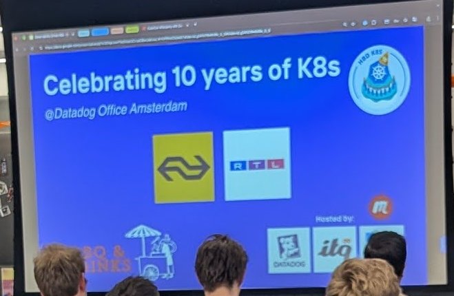 The Celebrating 10 years of K8s at Datadog Office Amsterdam title slide on the projector, with a Hosted by strip including the Datadog logo