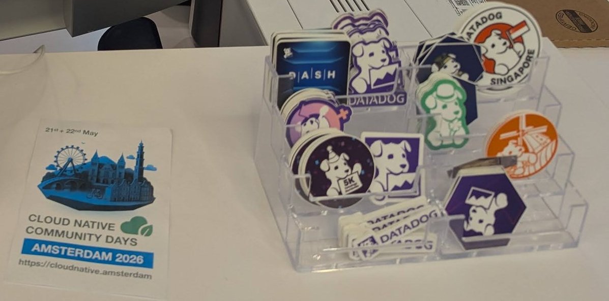 A flyer for Cloud Native Community Days Amsterdam 2026 on 21 and 22 May next to a rack of Datadog stickers