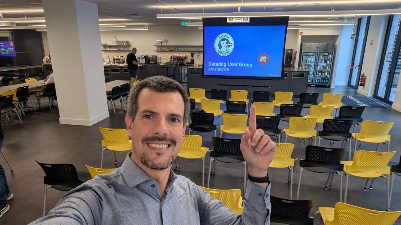 Luca Berton taking a selfie at the Datadog office in Amsterdam, with the Datadog User Group Amsterdam opening slide on the projector and empty yellow and black chairs