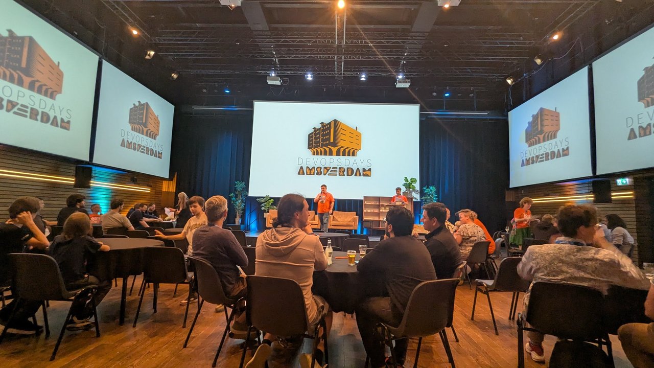 The DevOpsDays Amsterdam main hall set up with round tables and the event logo on the screens before the evening programme