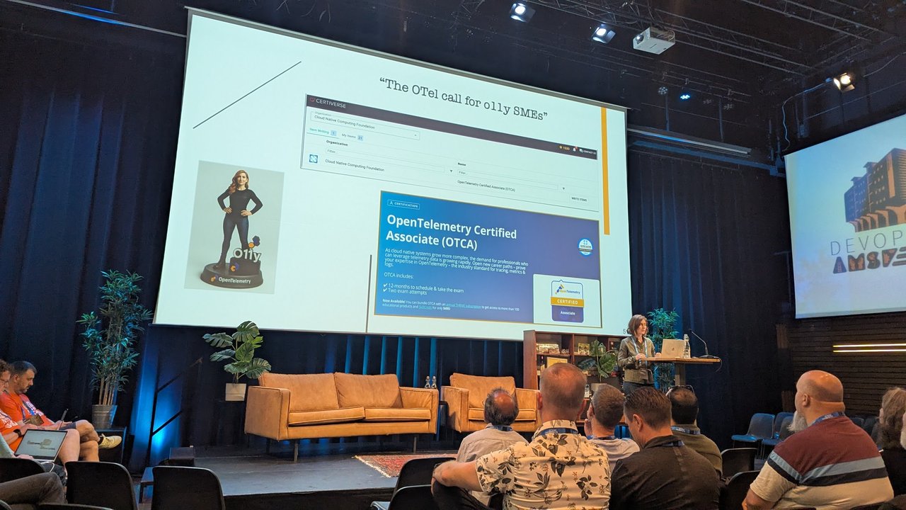 Speaker on stage at DevOpsDays Amsterdam 2025 beside a slide titled The OTel call for o11y SMEs, showing the OpenTelemetry Certified Associate certification