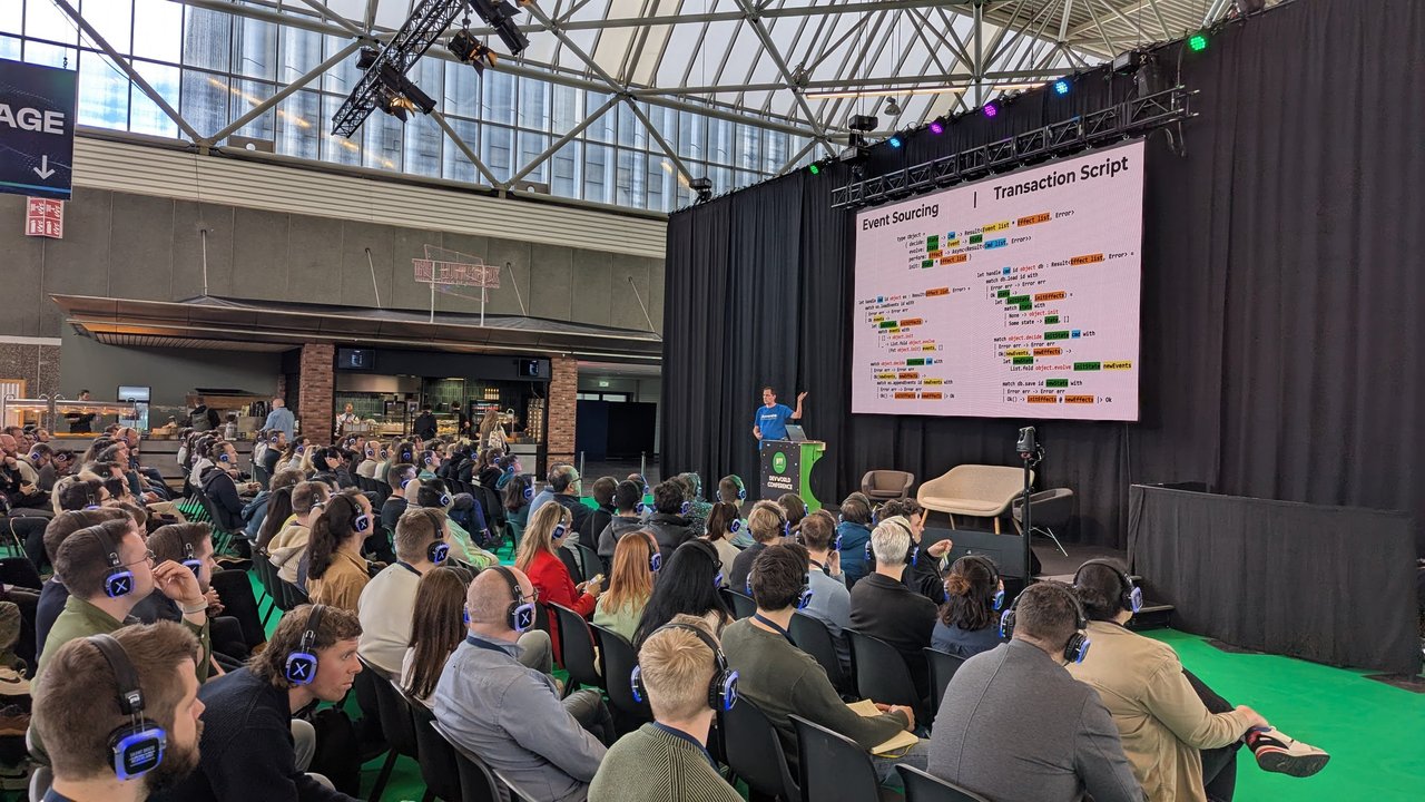 DevWorld 2025 slide comparing Event Sourcing and Transaction Script handlers in F#, with the speaker at the lectern