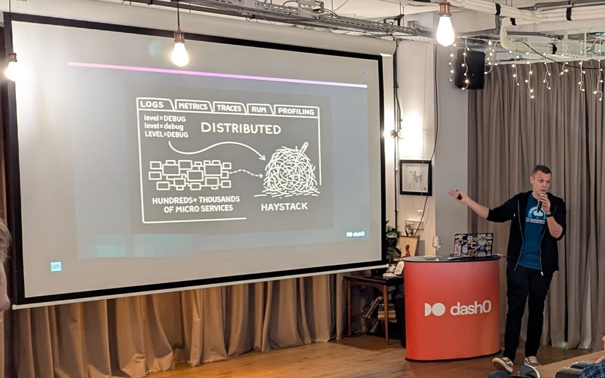 Kasper Borg Nissen pointing at a hand-drawn slide with tabs for logs, metrics, traces, RUM and profiling, showing hundreds or thousands of microservices feeding a haystack and three different spellings of level=DEBUG