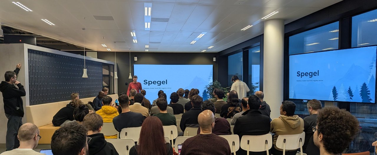 Audience in rows of chairs facing two screens that both show the Spegel title slide, Stateless cluster local OCI registry mirror, with the speaker standing to the left