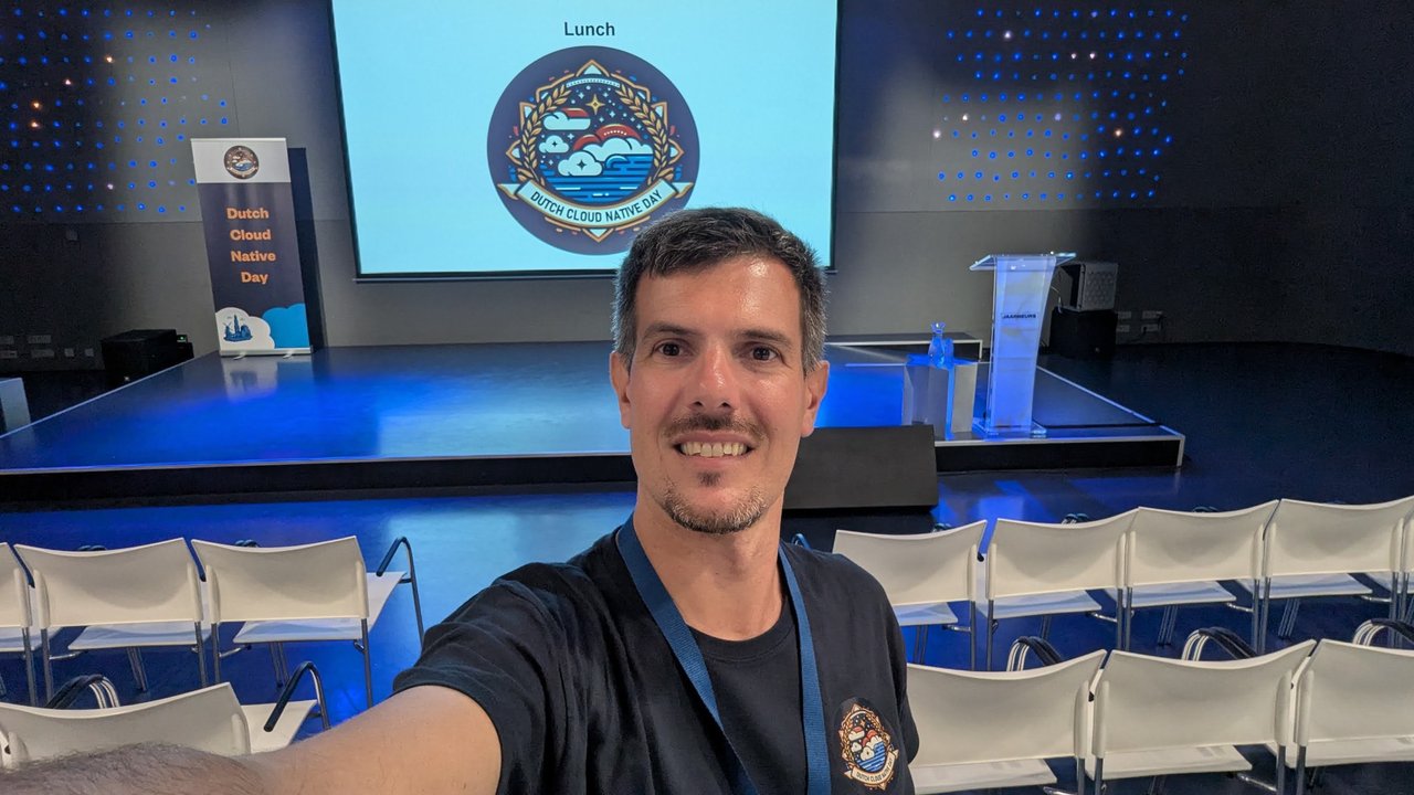 Luca Berton taking a selfie in front of the empty main stage at Dutch Cloud Native Day, with the lunch slide and the event badge logo on the screen