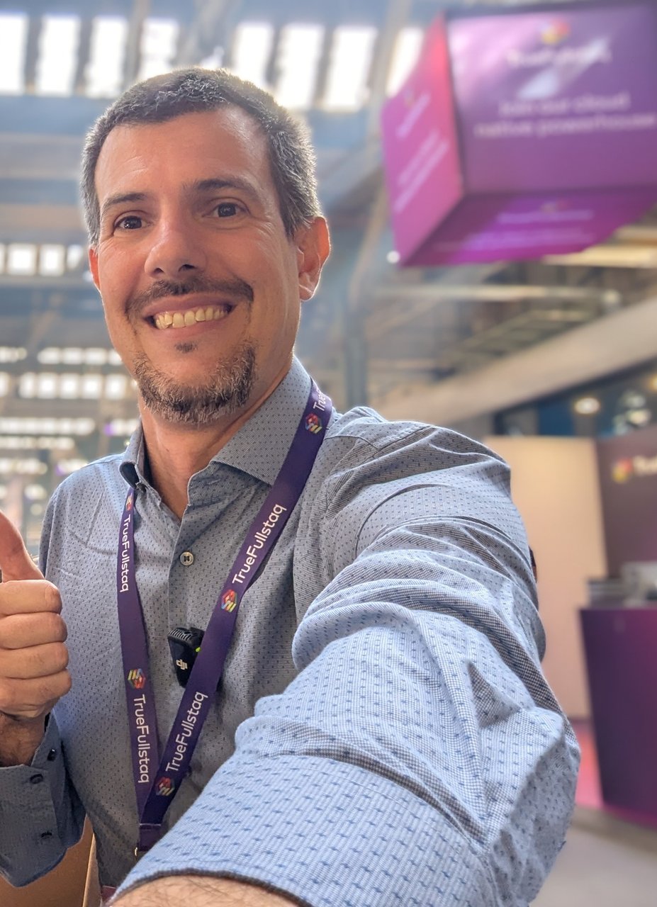 Selfie of Luca Berton with a TrueFullstaq lanyard on the EDGECASE 2025 expo floor, a purple TrueFullstaq cube hanging from the roof of the old carriage shed behind him