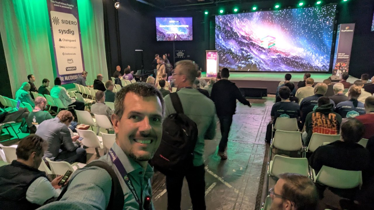 Selfie of Luca Berton in the EDGECASE 2025 main hall at the Rijtuigenloods in Amersfoort, rows of chairs filling up in front of a large LED wall showing a galaxy, and a banner listing the sponsors Sidero, Sysdig, Chainguard, Dell Technologies, Grafana Labs, AWS and Veeam under the title The Multiverse Saga
