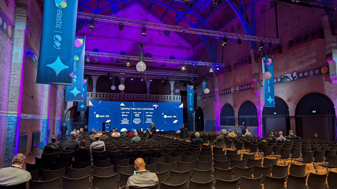 The ElasticON Amsterdam lightning-talk hall at the Beurs van Berlage, with brick arches, purple lighting, Elastic ON banners and the Lightning Talks over Lunch Break screen