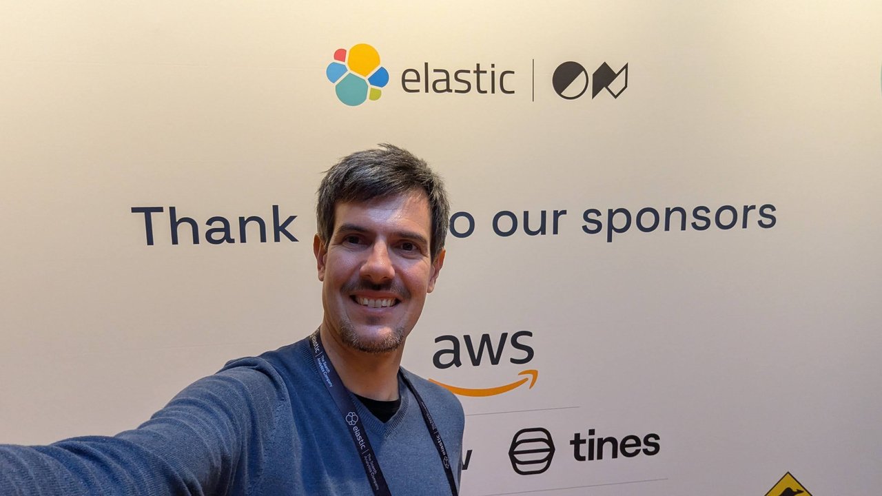 Luca Berton taking a selfie in front of the ElasticON Thank you to our sponsors wall with the AWS and Tines logos