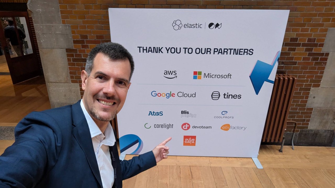 Luca Berton pointing at the ElasticON Thank you to our partners wall with AWS, Microsoft, Google Cloud, Tines, Atos, Corelight and Devoteam logos