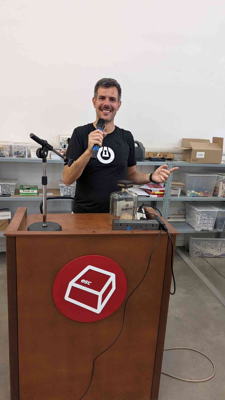 On stage at the ESC podium, microphone in hand — the wooden podium with the red ESC cube logo, Disaronno bottle, and power strips tell their own story